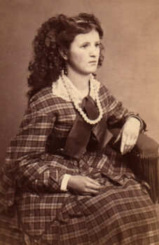A young woman with long brown hair, a plaid full dress with a lace collar, dark ribbon and pearls, sits in a chair. 