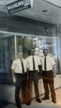 A black and white photo of an older man with glasses and two younger men, all wearing button-downed shirt sleeve shirts, dark ties and dark pants, in front of a store with a sign for "Wilhelms Wall Covering & Paint) above them and paint behind them.