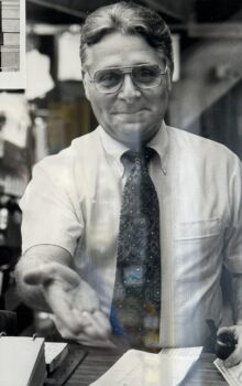 Black and white phot of a man with glasses, a short white button-down shirt and a dark tie, reaching out his hand.