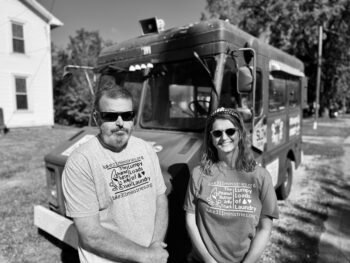 A man with short hair and a beard stands next to his wife with long hair and a smile in front of a large truck. 