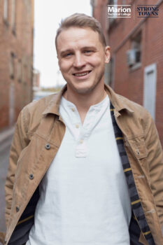 A white man with short light brown hair and a smile wears a white shirt and a tan jacket.