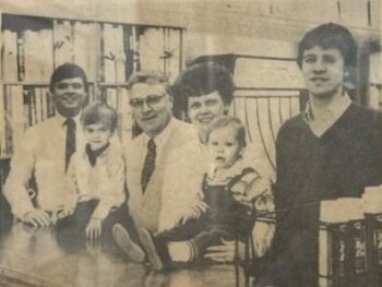 A black and white clipping, now yellowed, of two men in suits and ties, one in a dark shirt, a woman for with a white shirt, and two toddler boys surrounded by paint and wallpaper supplies at Wilhelms.