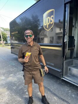 A UPS driver in uniform stands next to his truck and smiles while holding a treat.