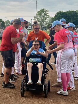 An unstoppable player in a wheelchair smiles while being pushed around the field by a woman and high fived by players of the opposing team wearing uniforms.