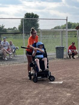 An Unstoppable player in a wheelchair holds a bat while wearing a helmet. An adult volunteer assists him at home plate.