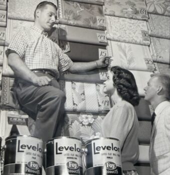 black and white Wilhelms ad from the 1960s showing a man showing wallpaper while standing behind paint cans to a couple of customers.
