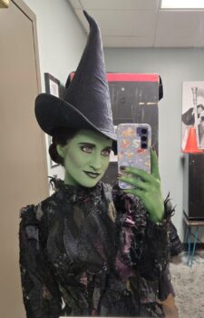 Carly in costume as Elphaba with green skin, a black witch's hat and black outfit. 