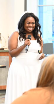 A Forge competitor wearing a white wedding dress talks to the judges at the front of an auditorium.