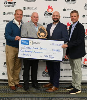 The donor, the president of the college, and the father in son who won the Forge competittion and trophy smile and hold a giant check and a wooden trophy.