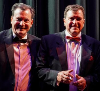 Two men dreessed white shirts, dark suits and bow ties smile.
