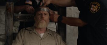 An older man with a white beard looks thoughtful while being connected to the electric chair by prison guards.