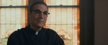 A man with short brown hair and black-rimmed glasses and a pastoral collar sits in front of a stained glass window.