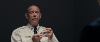 An older bald man in a police uniform holds a cigarette and lighter.