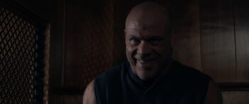 A blad man with stubble smiles with a wild look in his eyes inside a confessional booth.