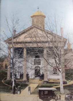 A colorized photo of a tall building with large windows, tall columns and a cupuloa. Men in suits and hats sand outside. There is a hrose-drawn carriage out front. 