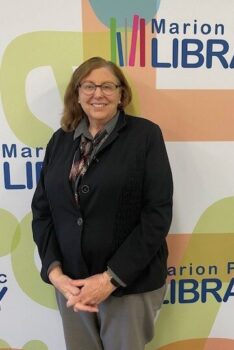 A white woman with shoulder-,ength wavy brown hair, a smile, black-rimmed glasses, pearl earrings, and a suit and scarf.